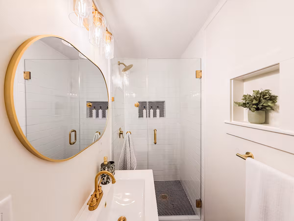 Bathroom with walk-in glass shower, gold fixtures, and round mirror
